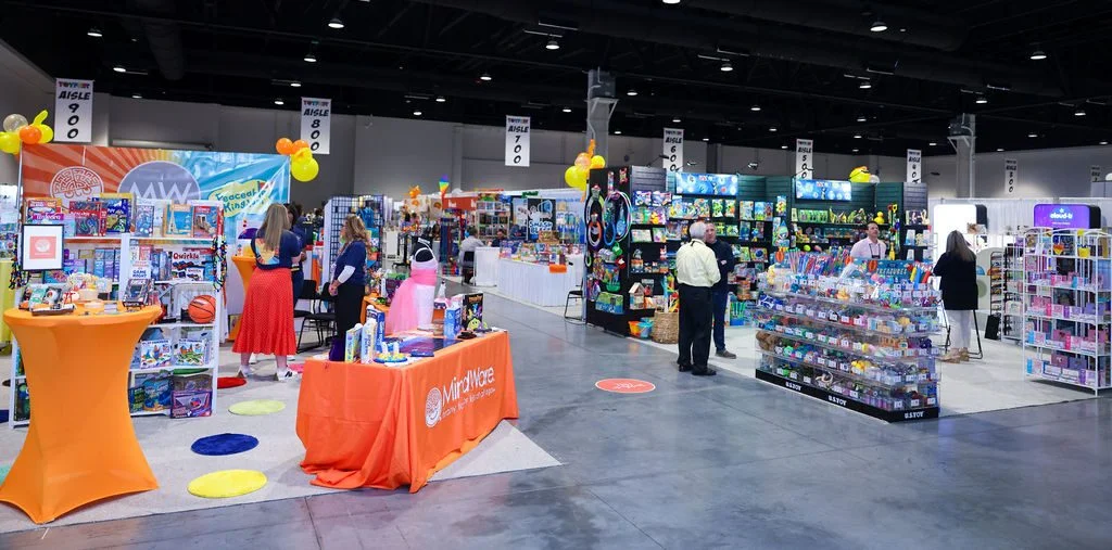 Colorful toy exhibition with various booths displaying games and products, crowded with visitors, featuring orange table and decorative balloons.